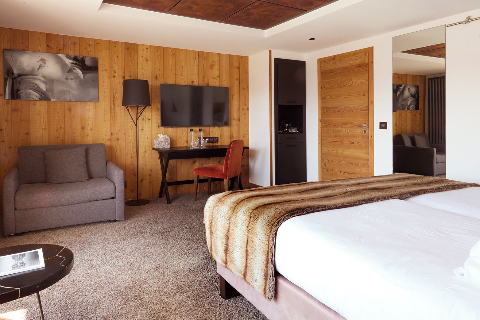 A hotel room with a bed, a sofa, a desk, a chair, a TV, and a painting on the wall.