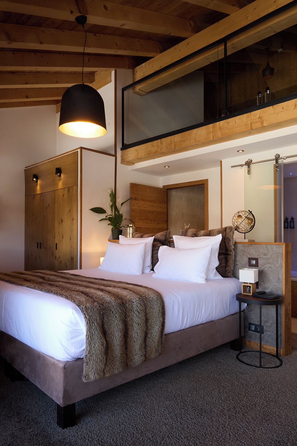 Une chambre avec un grand lit, des poutres apparentes en bois au plafond, un luminaire suspendu, une table de chevet avec une lampe et un miroir.