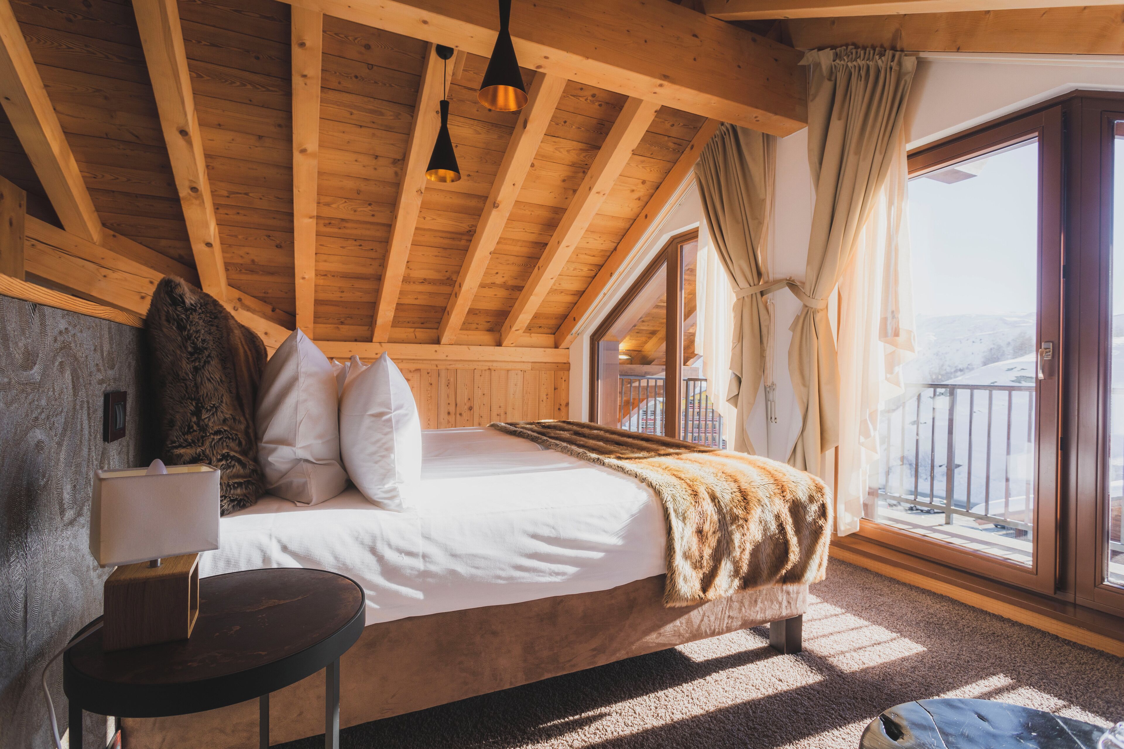 Une chambre avec un plafond en bois, un lit avec une literie blanche, un plaid en fourrure, une lampe de chevet et une vue sur l’extérieur.