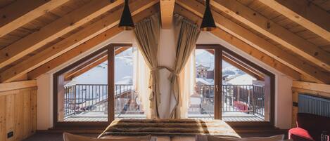 A high-ceilinged room with wooden beams, a large bed with fur throw pillows, and a view of a snowy mountain landscape through large windows.