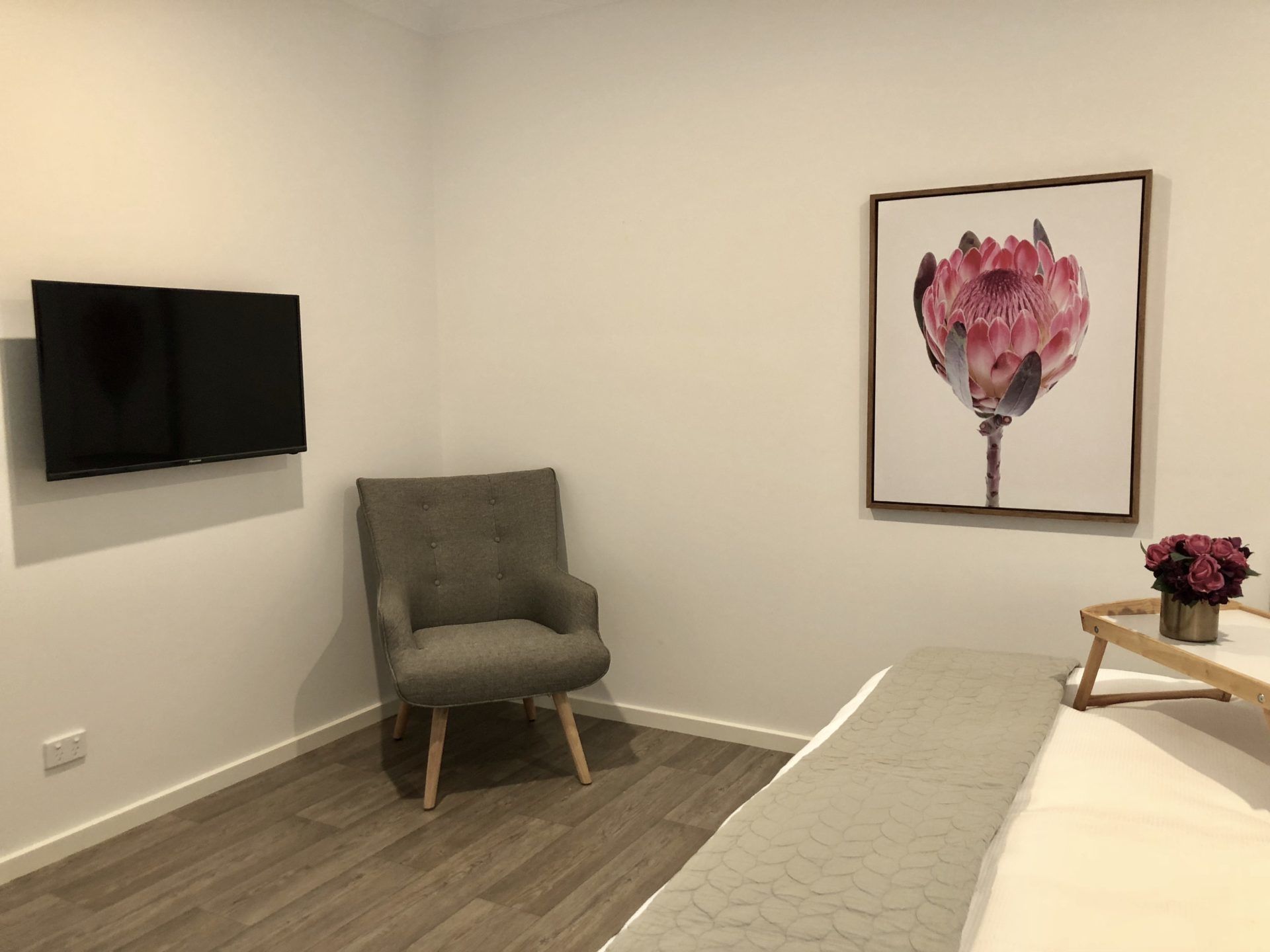 A room with a grey upholstered armchair, a flat-screen TV mounted on the wall, a framed floral artwork, a wooden side table with a vase of flowers, and a bed with a patterned bedspread.