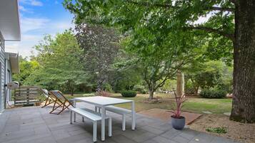 Um pátio externo com mesa e bancos, cercado por árvores e um jardim.