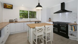 A modern kitchen with white cabinetry, a wooden countertop, a large window, a black range hood, and a built-in oven. - Marshmallow Retreat (Bright)