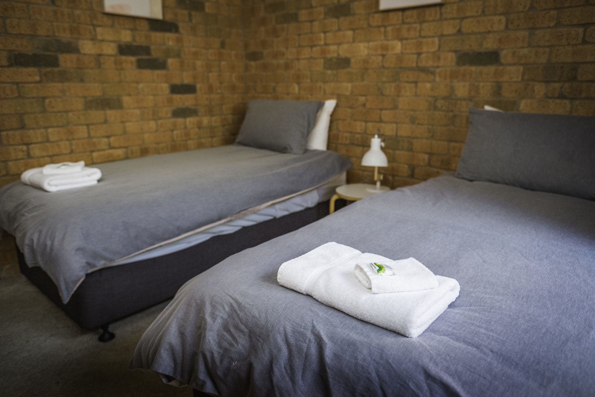 A hotel room with two beds, grey bedding, a brick wall, and a small bedside table with a lamp.