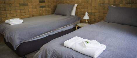 A hotel room with two beds, grey bedding, a brick wall, and a small bedside table with a lamp.