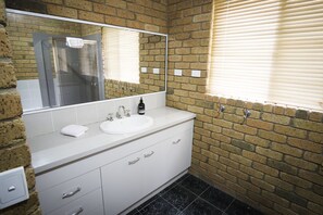 A bathroom with a white sink, a mirror, and a shower. The walls are made of brick, and there is a window with blinds.