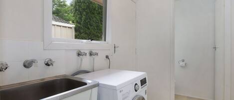 A laundry room with a washing machine, a sink, and a window overlooking a garden.