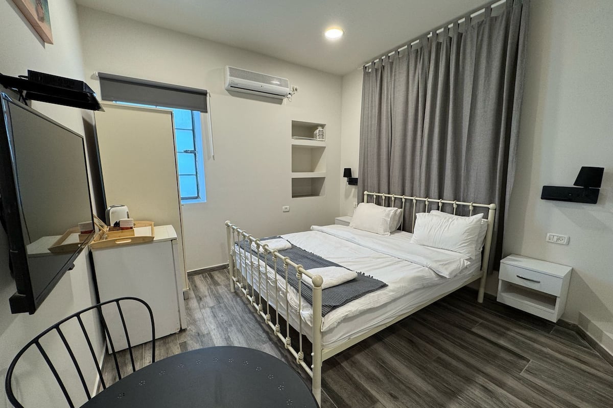 A compact hotel room with a single bed, a small desk, a chair, a mirror, and a window with curtains.