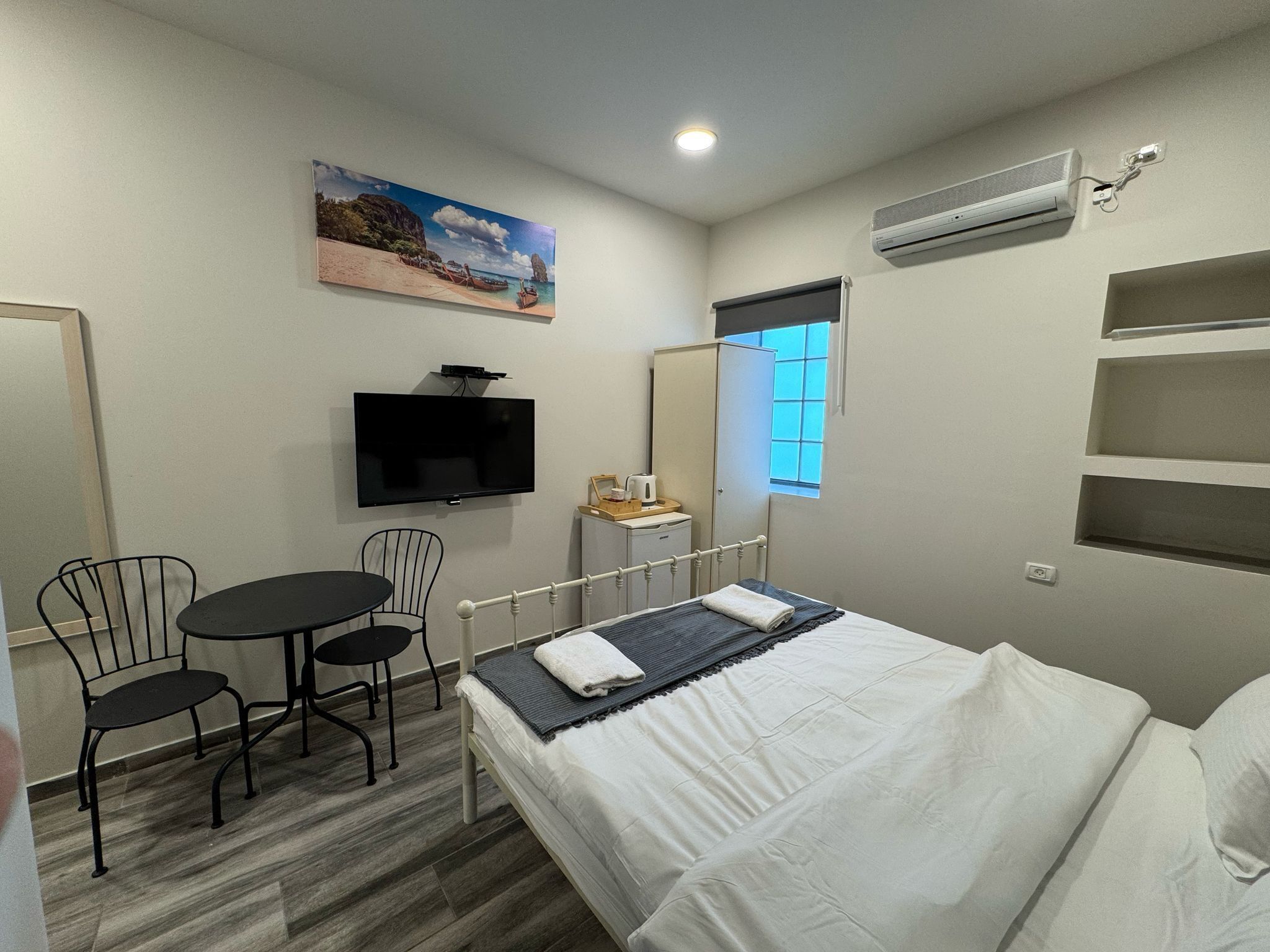 A bedroom with a bed, a desk with a chair, a television, a window, and an air conditioning unit.