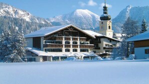 Um hotel com telhado coberto de neve, uma torre de sino e montanhas ao fundo.