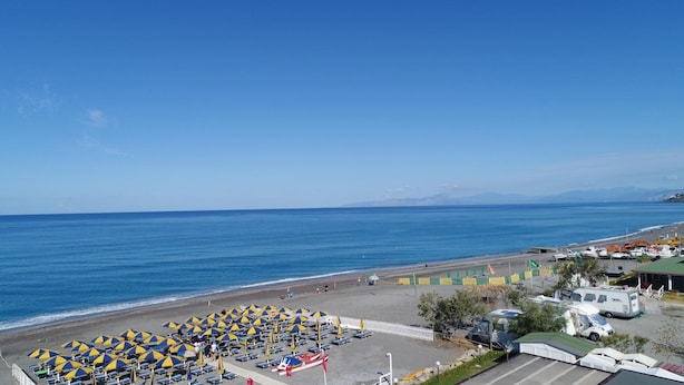 A beach with rows of umbrellas and sun loungers, a parking area with vehicles, and a clear view of the sea and distant mountains.