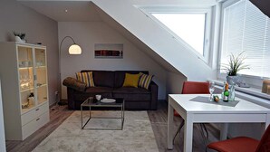 Un appartement moderne de style « loft » avec un plafond en pente, un canapé marron foncé agrémenté de coussins à rayures, une vitrine, une table à manger blanche avec une chaise rouge et une grande fenêtre équipée de stores.