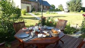 A wooden deck with a table set for breakfast, surrounded by lush greenery and a house in the background. - B&B L'Etape Normande & SPA (Montroty)