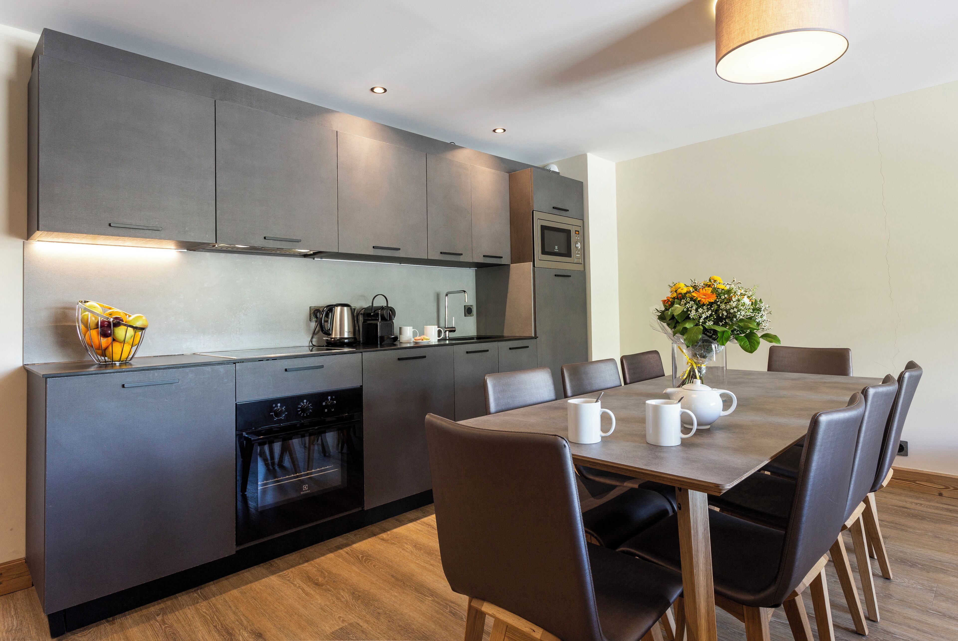 Una cocina moderna con armarios grises, horno empotrado y comedor con mesa y sillas.