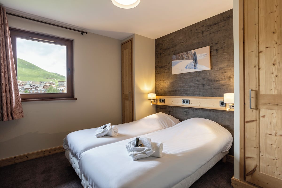 Ein Hotelzimmer mit zwei Betten, einem fenster mit Vorhängen, einem hölzernen Kopfteil, einer Wandleuchte und einem Gemälde.