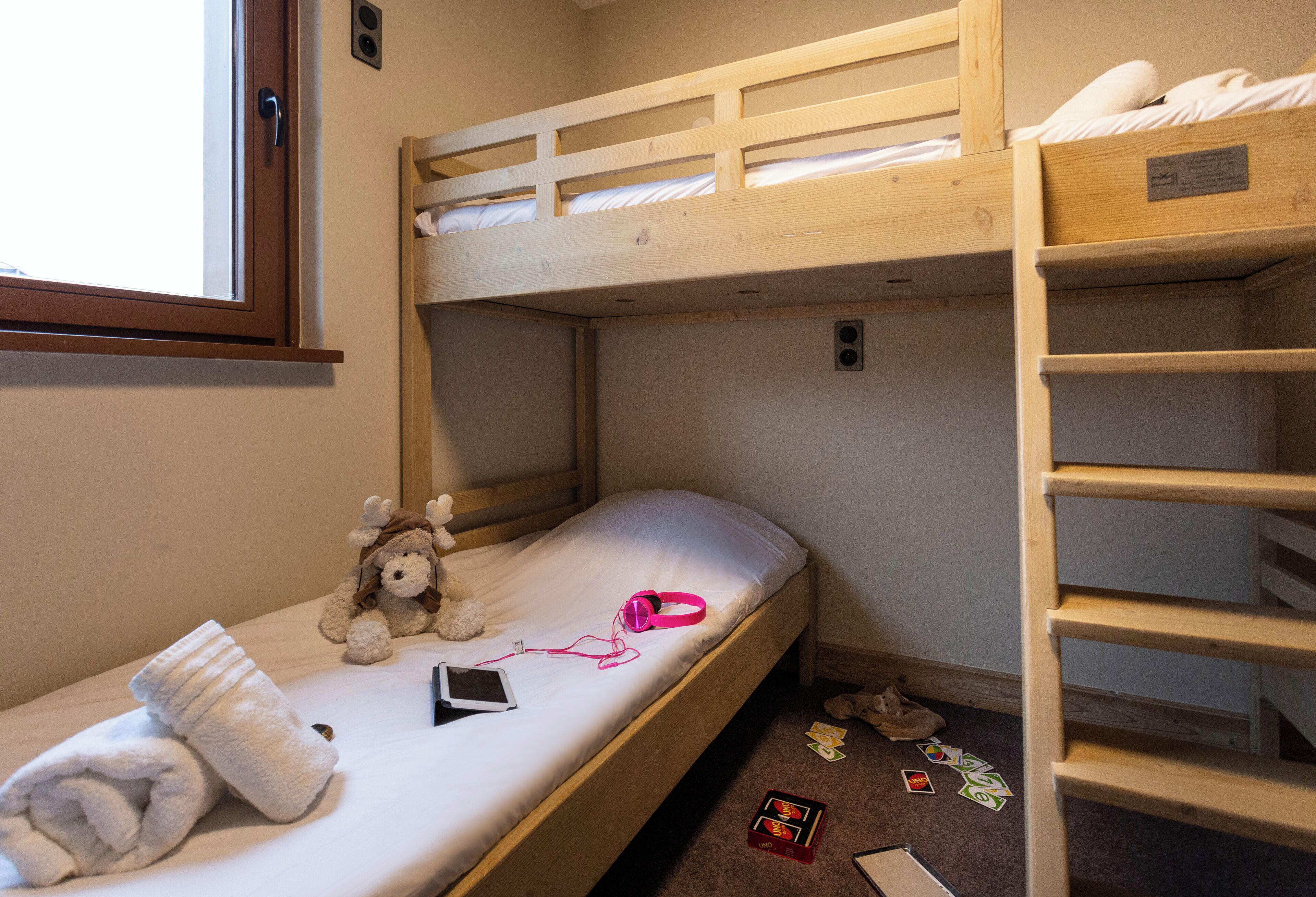 Habitación con literas, una ventana, un oso de peluche, audífonos y un teléfono inteligente en la litera inferior.