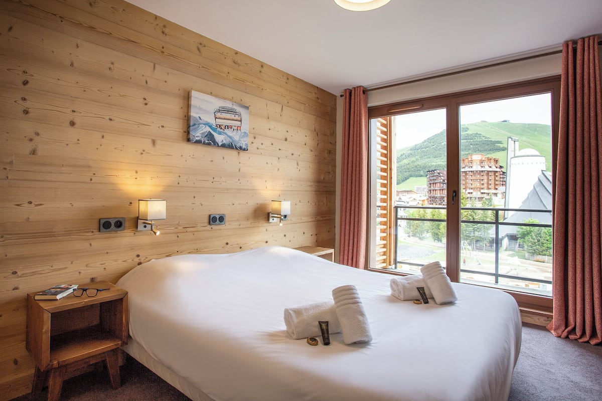 Ein Hotelzimmer mit einer Holzwand, einem großen Bett, zwei Nachttischlampen, einem Fenster mit Vorhängen und Blick auf Gebäude.