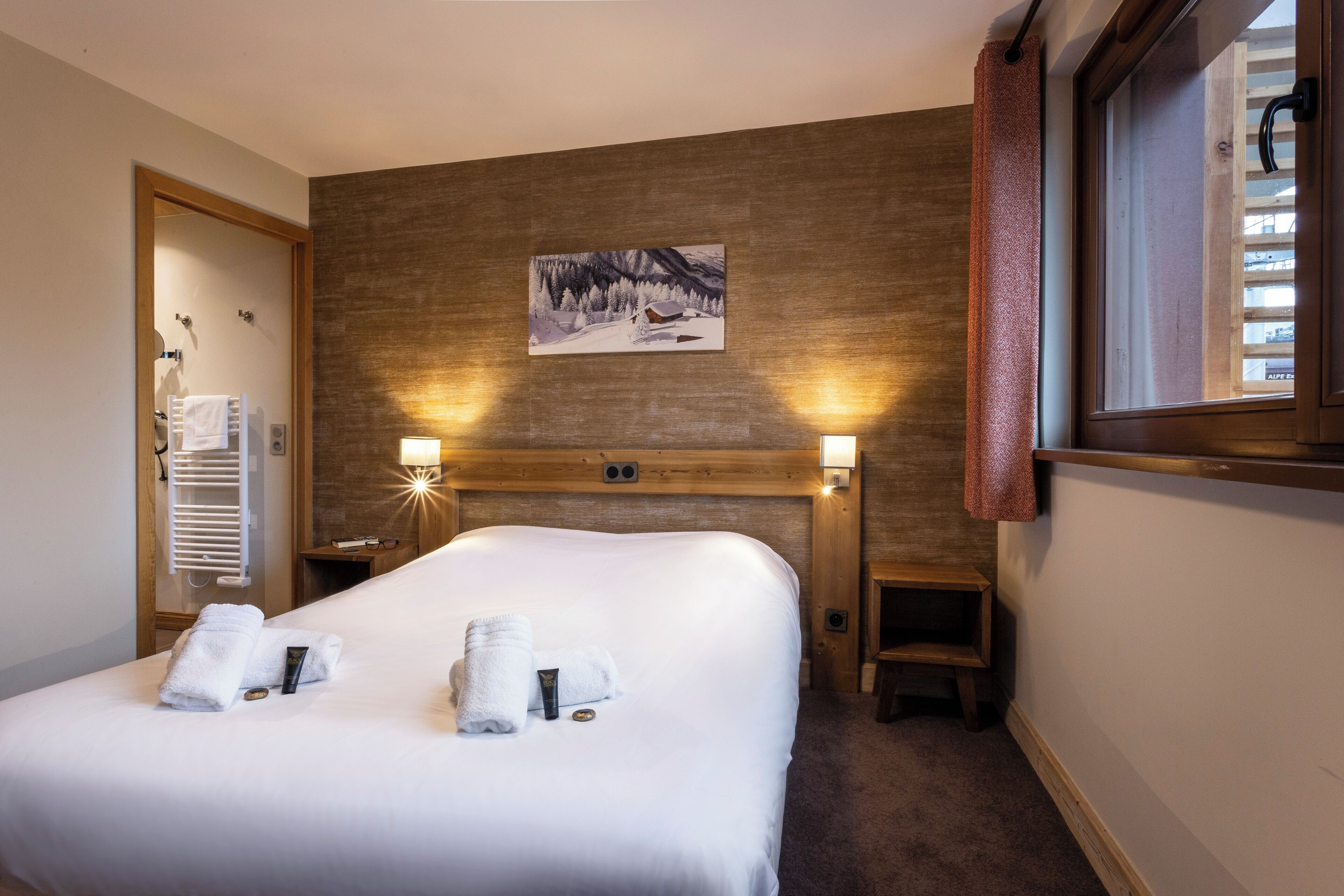Una camera d'albergo con un letto grande, due lampade sui comodini, un quadro appeso al muro, una finestra con tende e una testiera in legno.