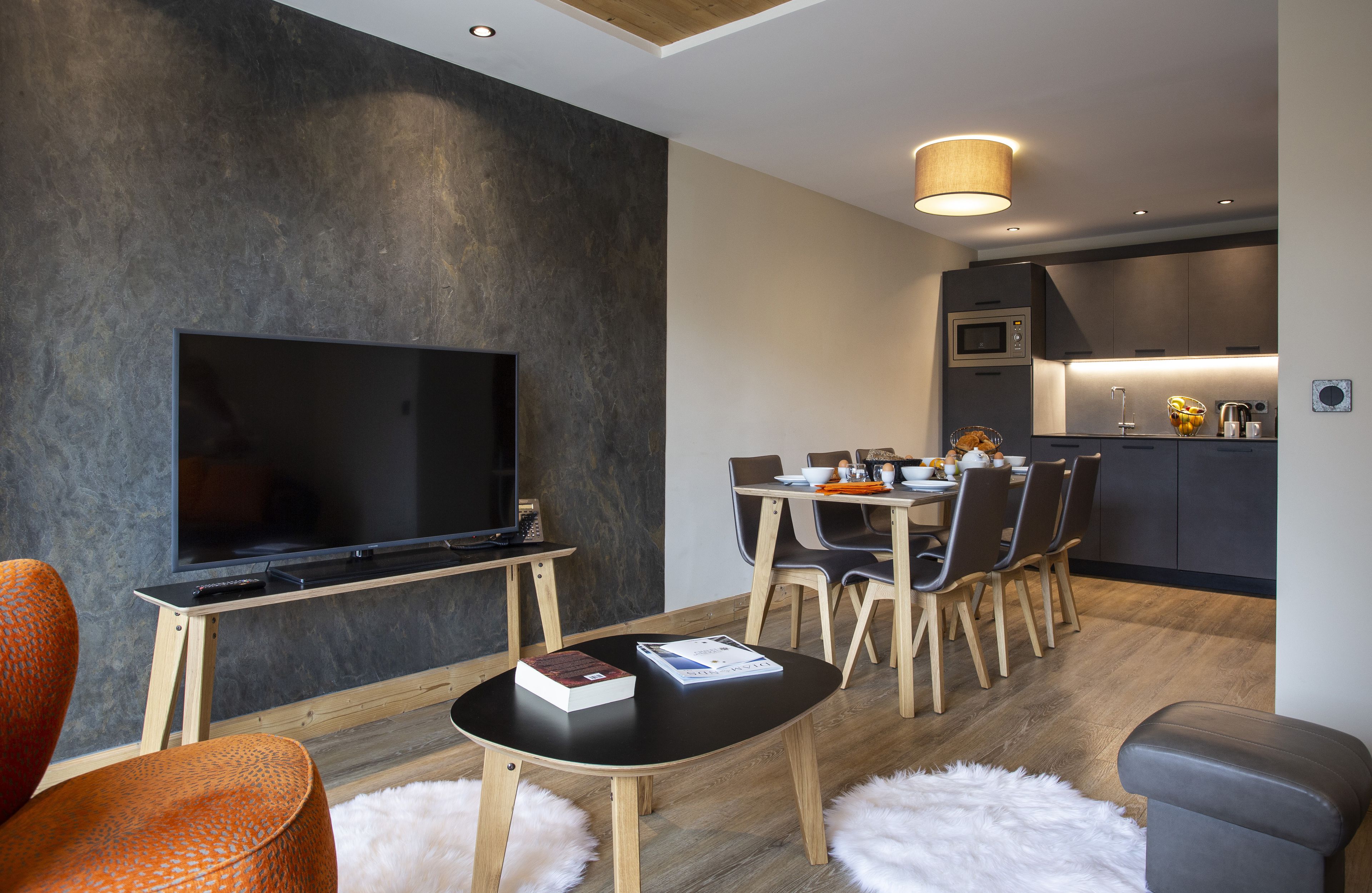 A modern living room with a flat-screen TV, a dining area with a wooden table and chairs, and a kitchen with a microwave.