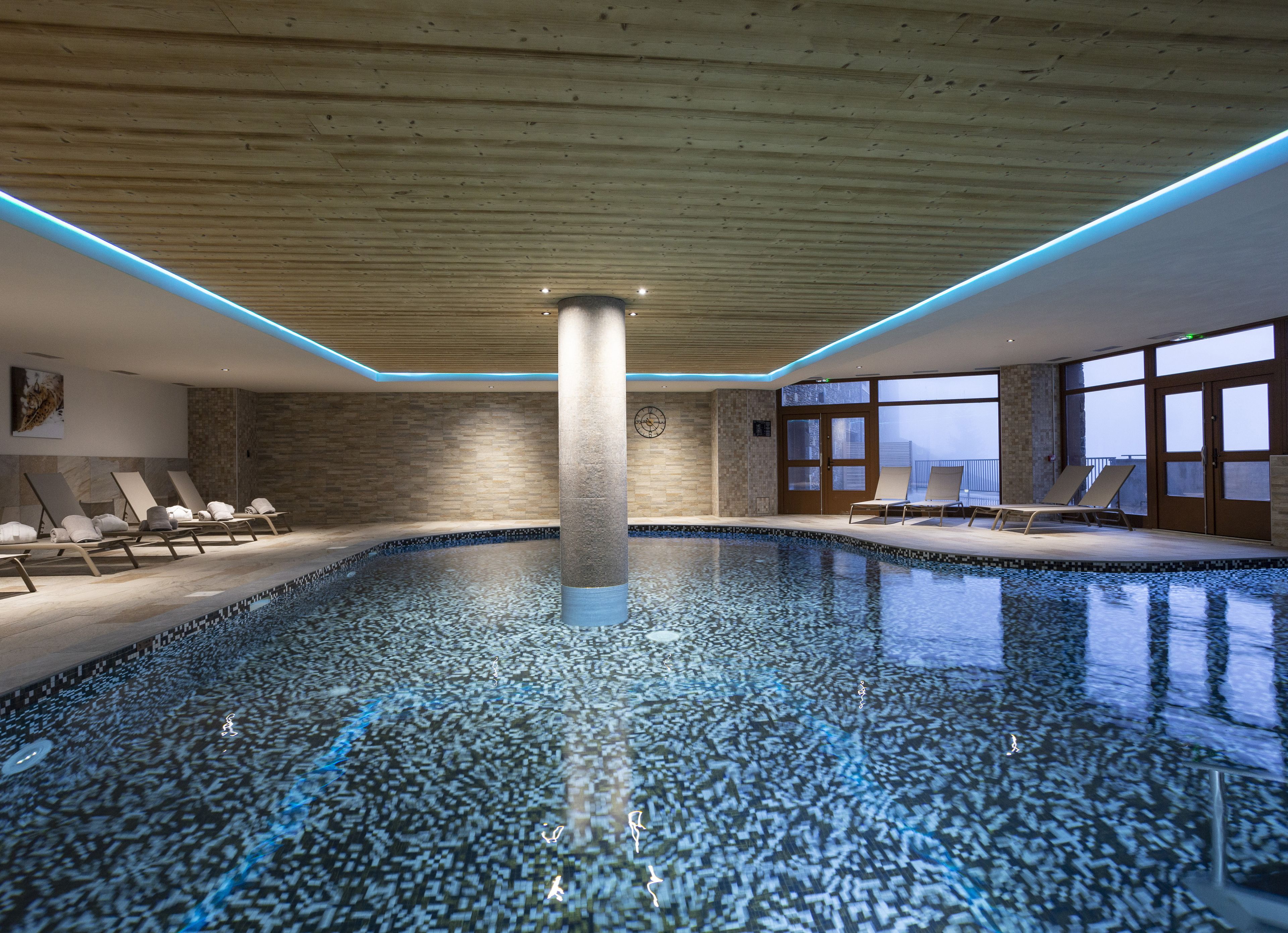 An indoor swimming pool with a wooden ceiling, lounge chairs, and large windows.
