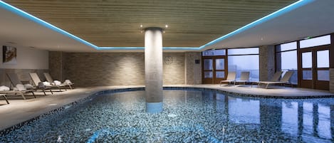 An indoor swimming pool with a wooden ceiling, lounge chairs, and large windows.