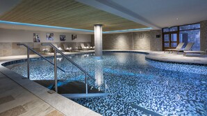 Une piscine intérieure dotée d’un plafond en bois, d’un mur en pierre et offrant une vue sur un bâtiment à travers de grandes fenêtres.
