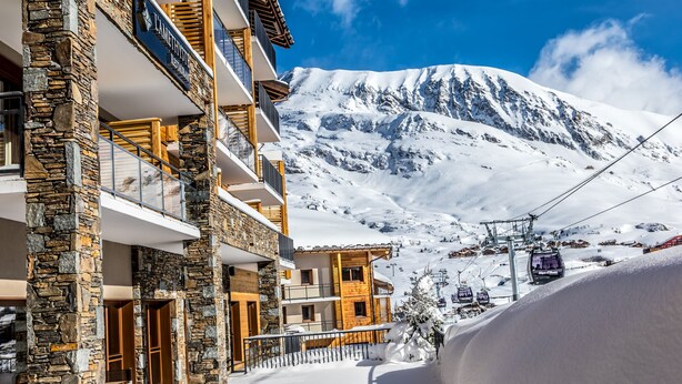 Un immeuble à plusieurs étages, avec une façade en pierre et en bois, un télésiège en arrière-plan et une montagne enneigée.