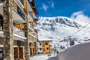 Un immeuble à plusieurs étages, avec une façade en pierre et en bois, un télésiège en arrière-plan et une montagne enneigée.