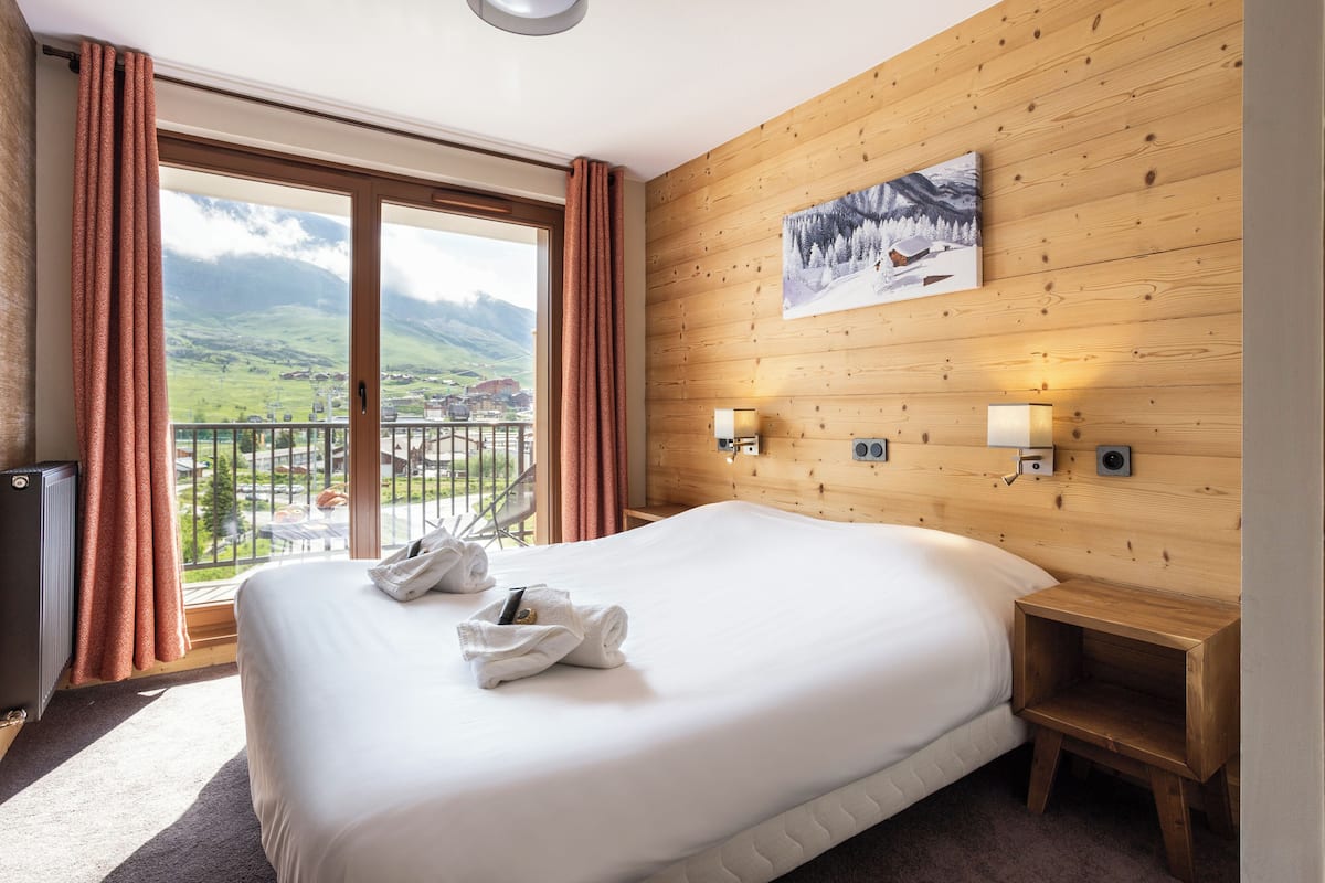 Ein Hotelzimmer mit einem großen Bett, Holzwandverkleidung, einem Gemälde, zwei Nachttischlampen, einem kleinen Nachttisch und Blick auf einen Balkon mit einer reizvollen Landschaft.