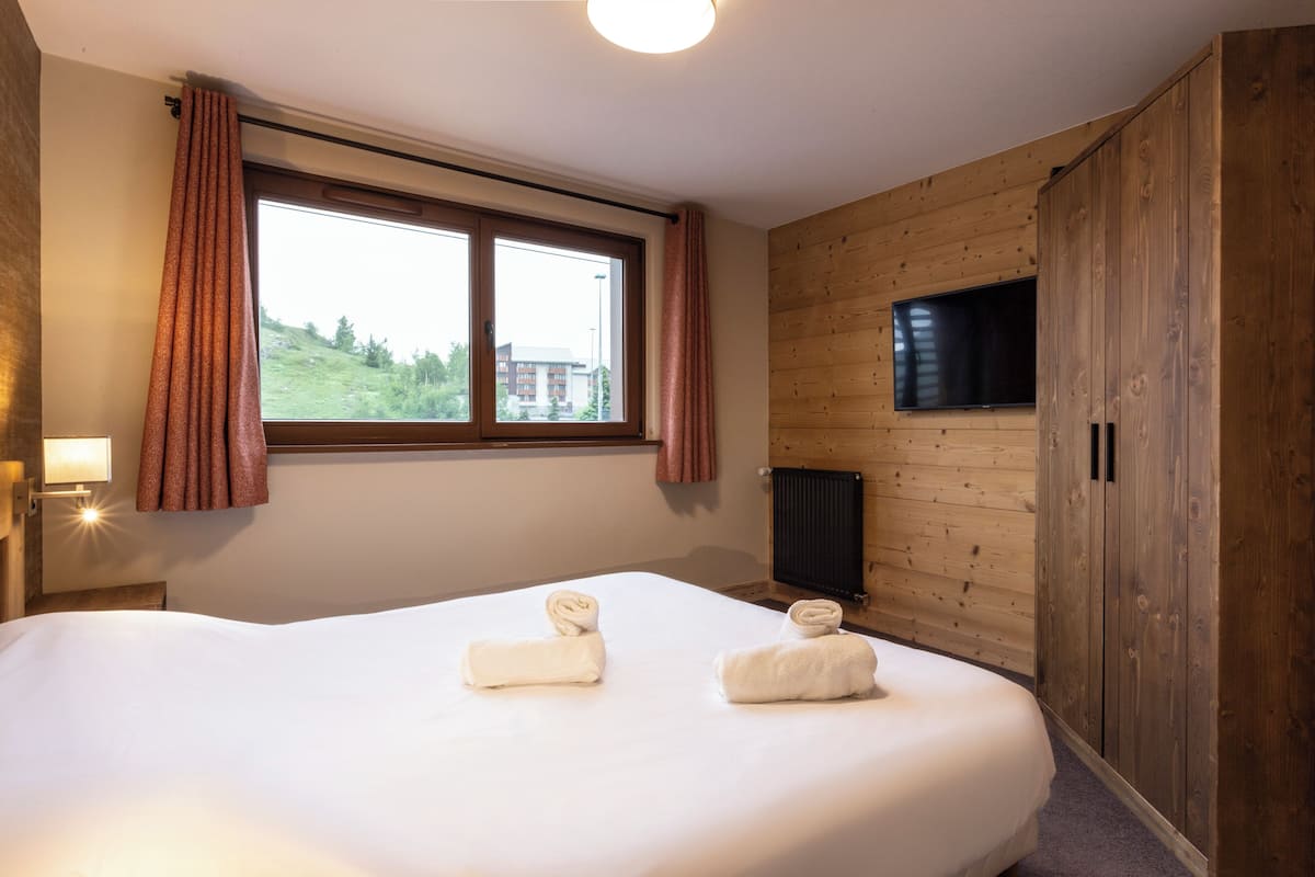 Ein Hotelzimmer mit einem großen Bett, zwei Handtüchern, einem fenster mit Vorhängen, einem Fernseher und Holzvertäfelung.
