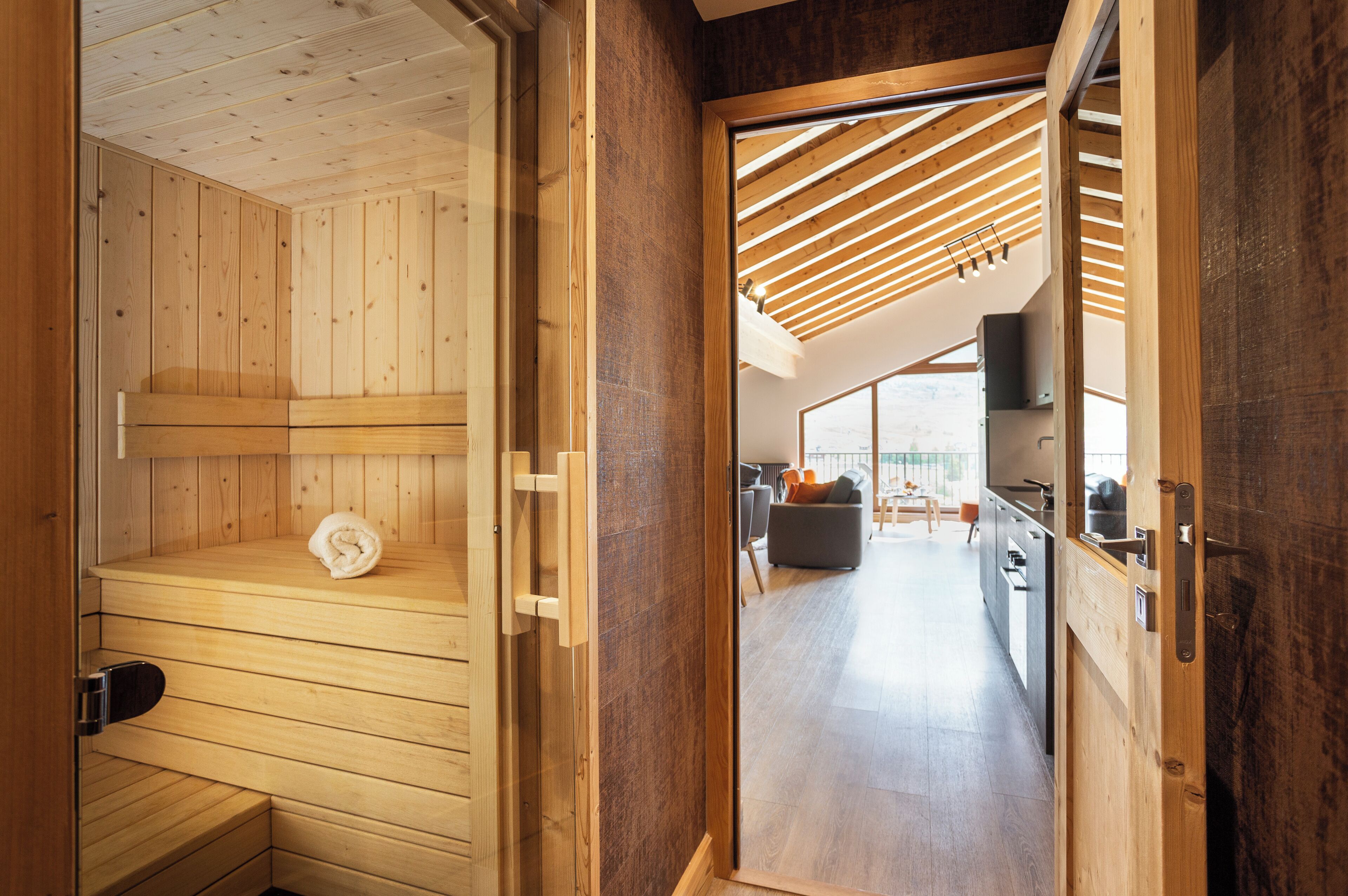 Un sauna de madera con banco y toalla, que da a una cocina moderna y zona de estar con techo inclinado y grandes ventanales.