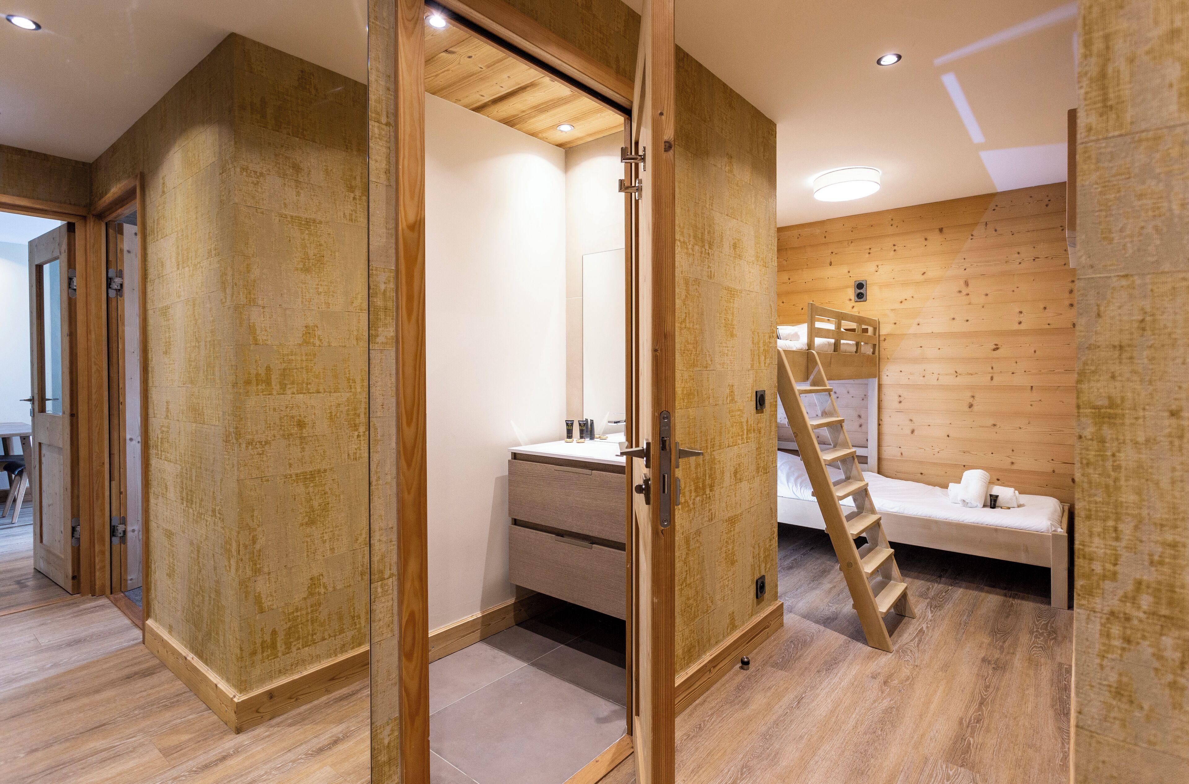 A room with a wooden floor, a bed with pillows, a bedside table, a mirror, and a wooden wardrobe.