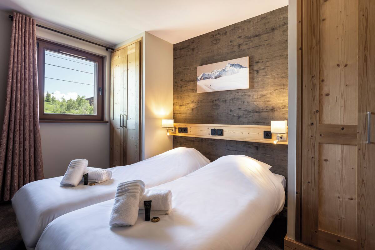Ein Hotelzimmer mit zwei Betten, einem fensterförmigen Durchbruch mit Vorhängen, einer Holzwand und einem aufgehängten Bild.