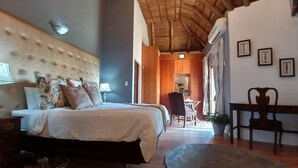 Un dormitorio con cabecera capitonada, cama con almohadas florales, mesita de noche y una silla. Se ve un área de comedor con una mesa y sillas a través de un umbral abierto.