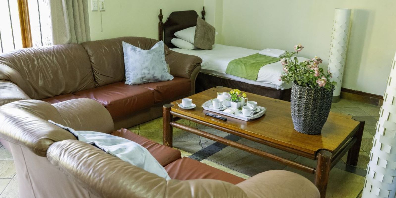 Een gezellige kamer met een bruine leren bank, een houten salontafel met een dienblad met kopjes en een vaas met bloemen, en een bed met een groene deken.
