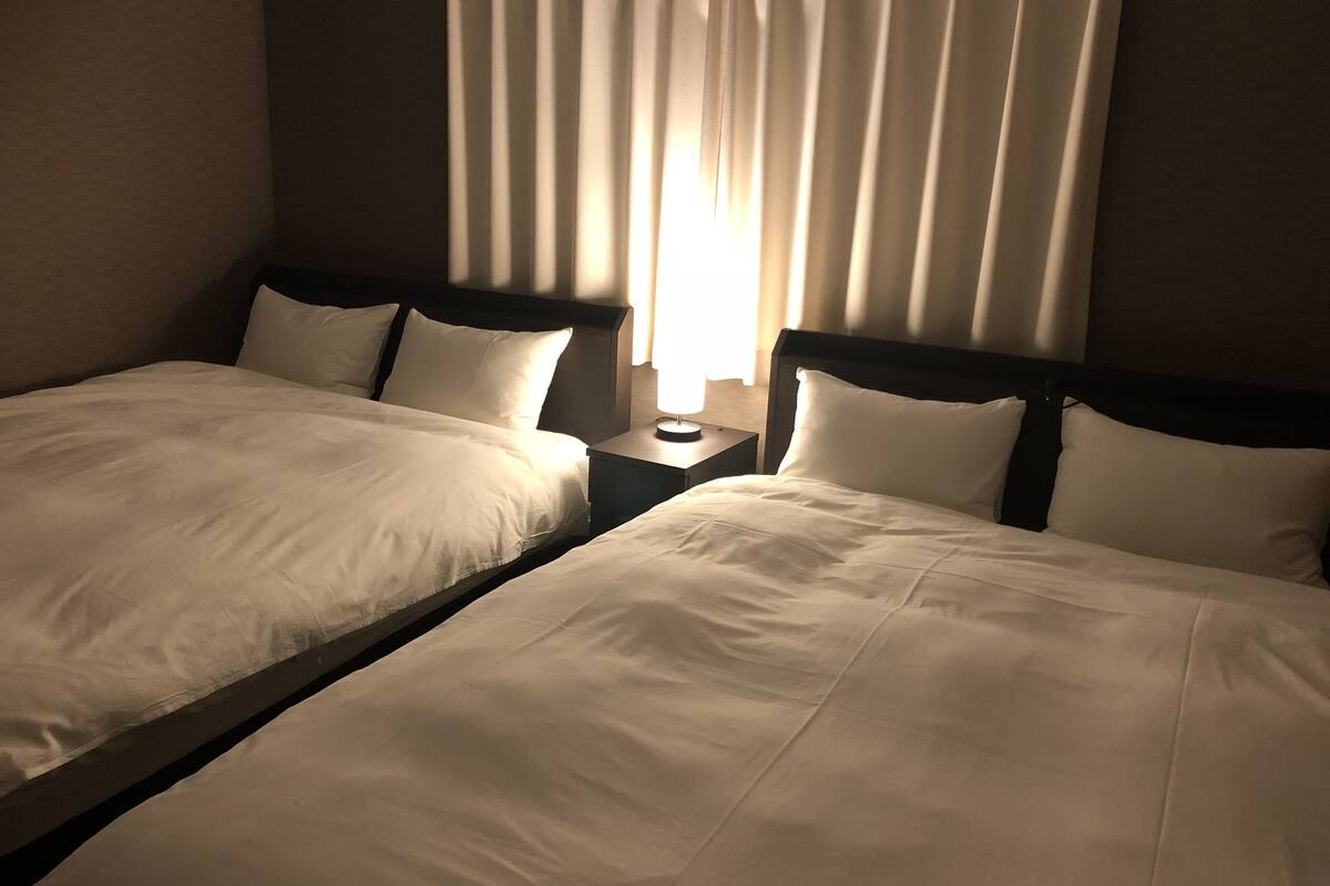 Dos camas individuales con sábanas blancas, una mesita de noche con lámpara y una ventana con cortinas.