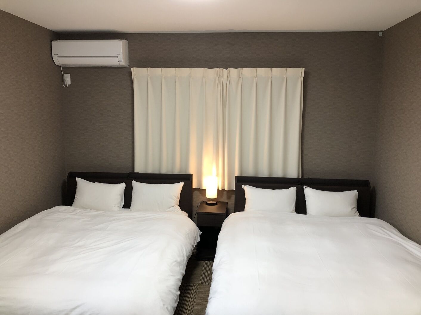 Ett hotellrum med två separata sängar, ett nattduksbord med en lampa, en väggmonterad luftkonditioneringsanläggning och ett fönster med gardiner.