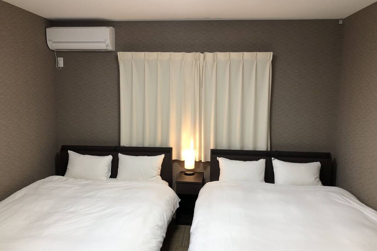Habitación de hotel con dos camas individuales, una mesita de noche con lámpara, aire acondicionado instalado en la pared y una ventana con cortinas.