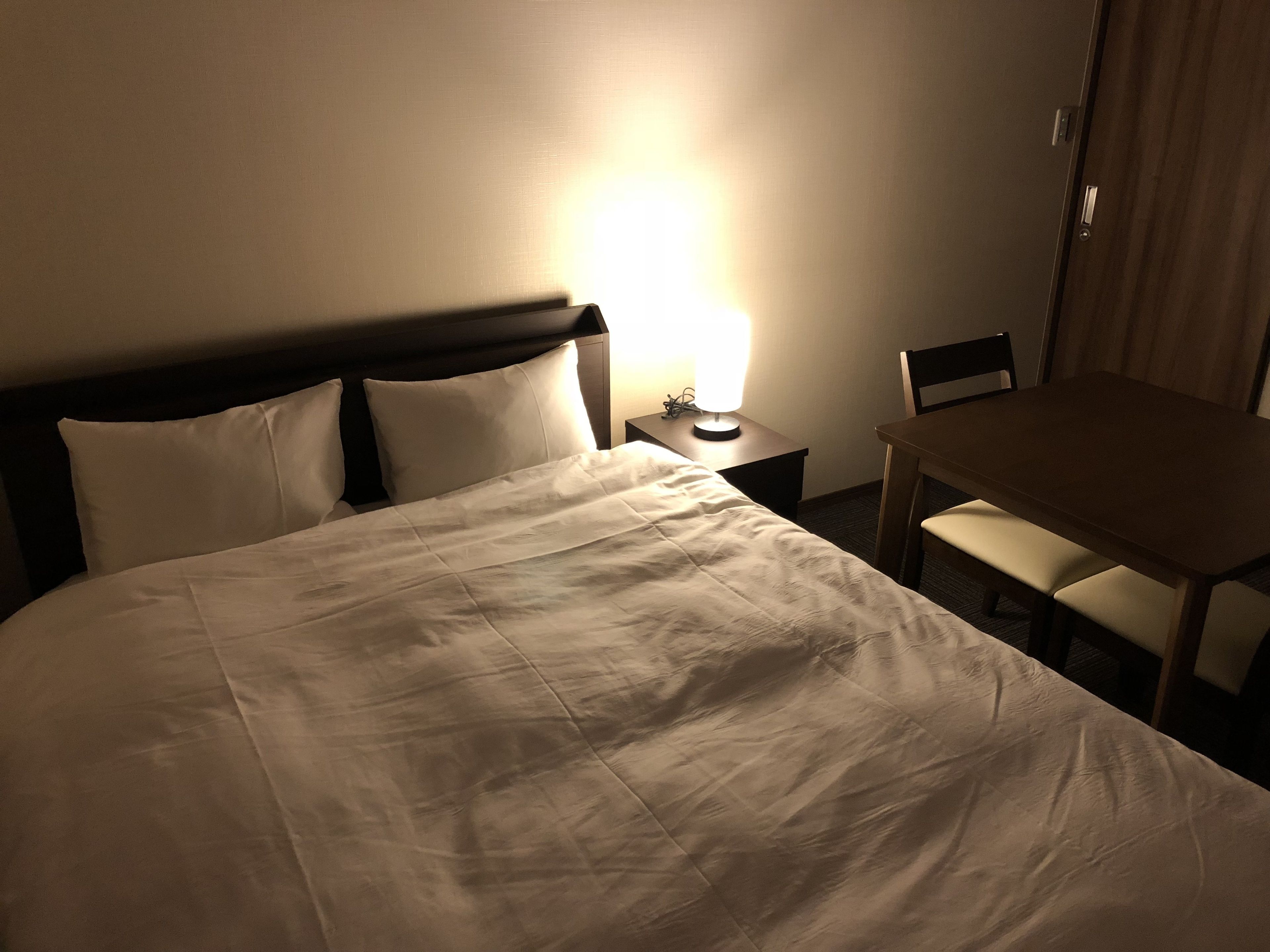 A hotel room with a large bed, a bedside table with a lamp, a wooden chair, and a small table.