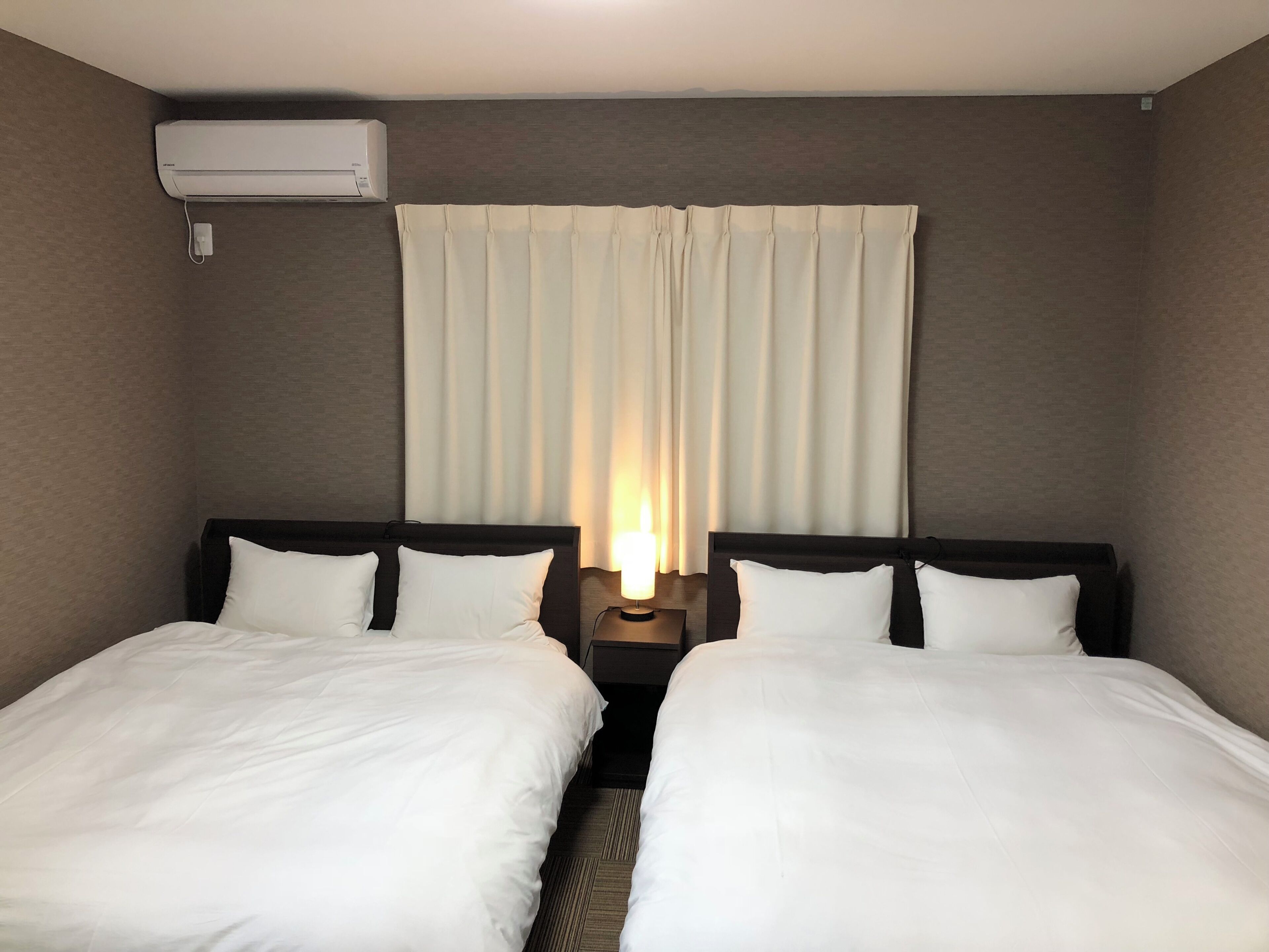 A hotel room with two beds, a nightstand with a lamp, a wall-mounted air conditioner, and a window with curtains.