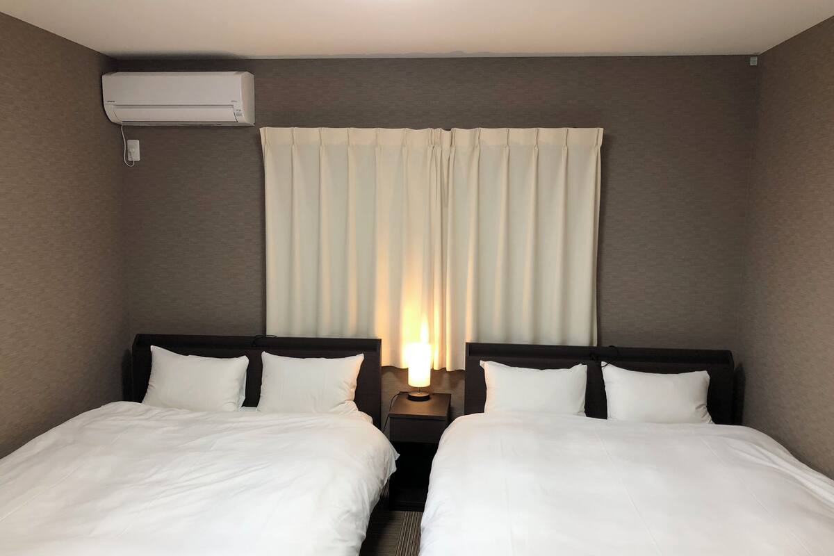 Habitación de hotel con dos camas, una mesita de noche con lámpara, aire acondicionado instalado en la pared y una ventana con cortinas.