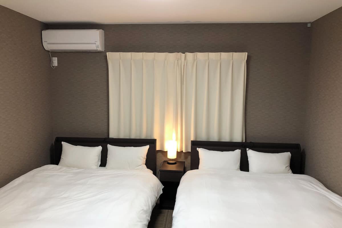 Une chambre d’hôtel avec deux lits, une table de chevet avec une lampe, un climatiseur fixé au mur et une fenêtre avec des rideaux.