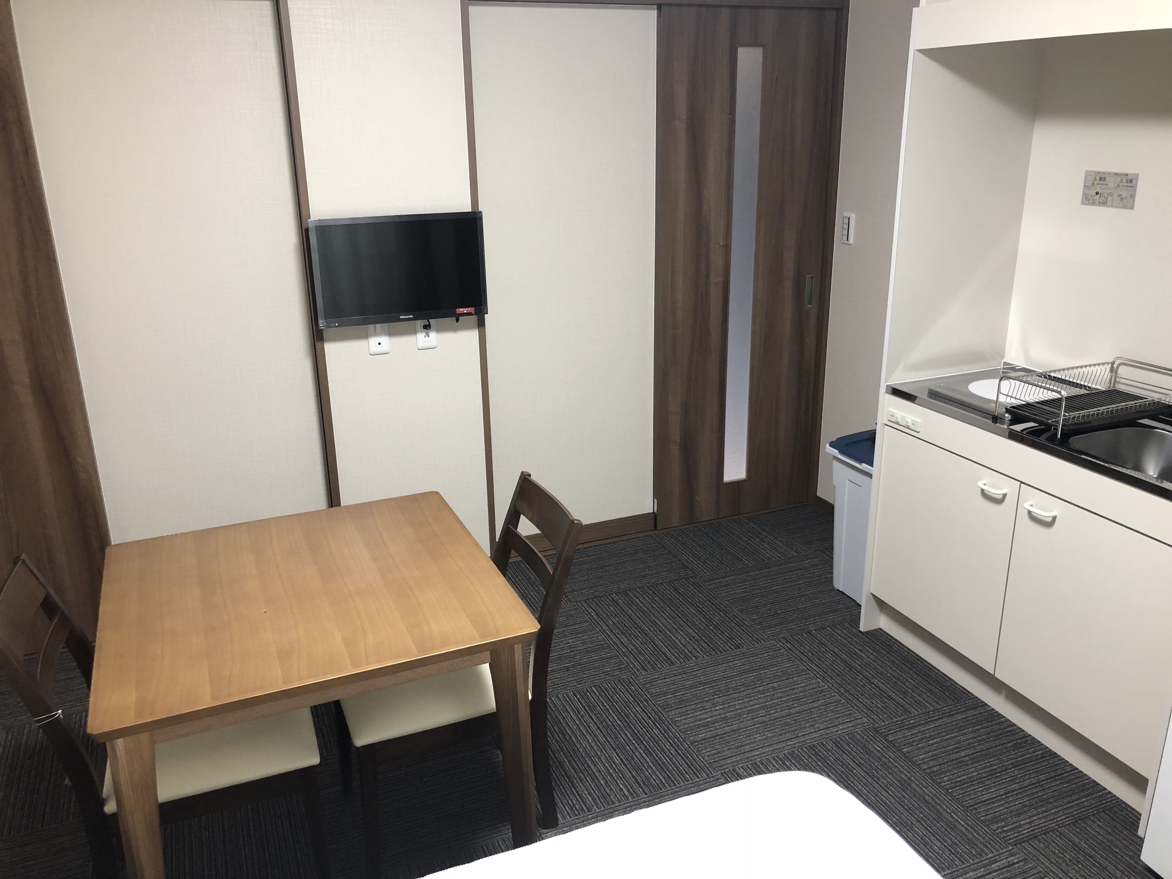 Ein kleines Zimmer mit einem Holztisch, einem einzelnen Stuhl, einer kleinen Küchenzeile mit Spüle und Mikrowelle sowie einem an der Wand befestigten Fernseher.