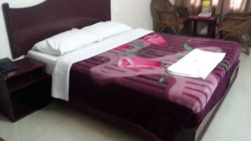 Uma cama bem arrumada com almofada branca e colcha floral roxa. Tem uma mesinha de cabeceira de madeira com uma luminária ao lado da cama.