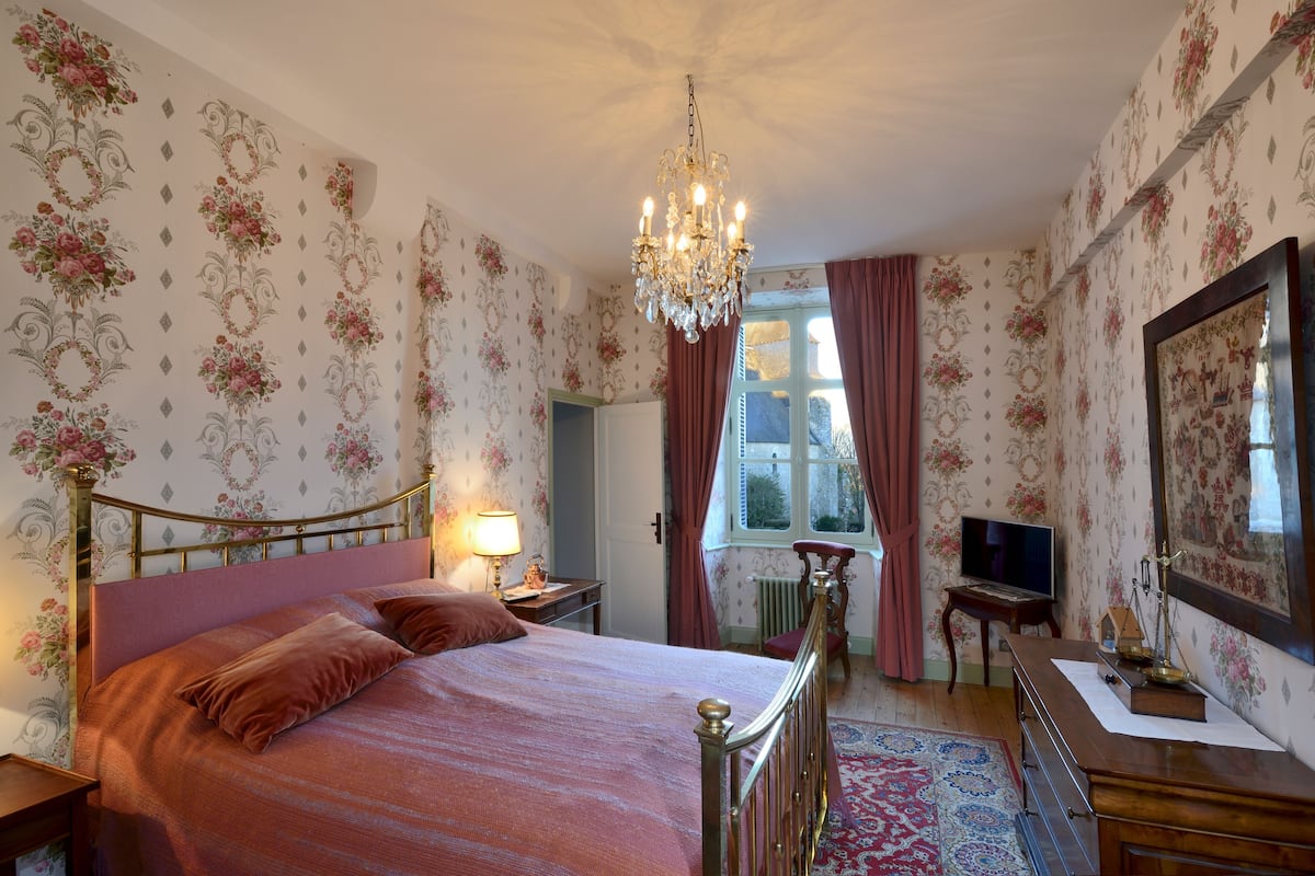 A bedroom with floral wallpaper, a chandelier, a bed with a headboard, a dresser, a mirror, and a window with curtains.
