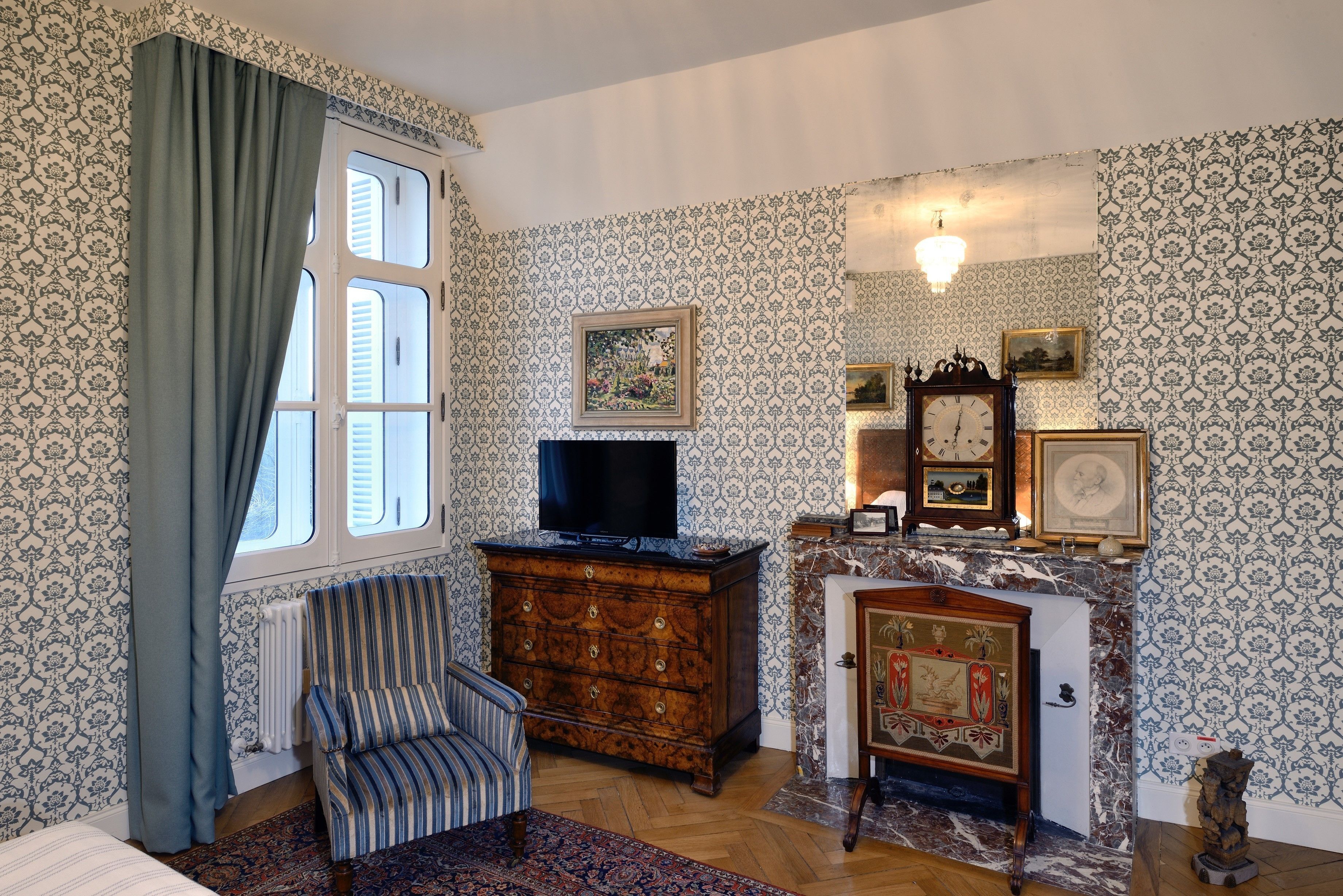 A traditional room with patterned wallpaper, a wooden dresser, a television, a fireplace with a clock, and a striped armchair.