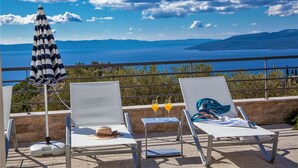 Two lounge chairs with a view of the sea and mountains, a striped umbrella, and two drinks on a table.