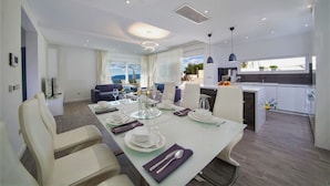 A modern dining area with a long table set for four, a kitchen area, and a seating area with a view.
