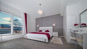 A modern bedroom with a large bed, bedside tables, a mirror, and a view of the sea.