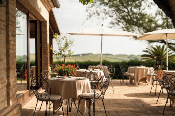 Un patio exterior con mesas y sillas, vista al campo y un edificio con grandes ventanales.